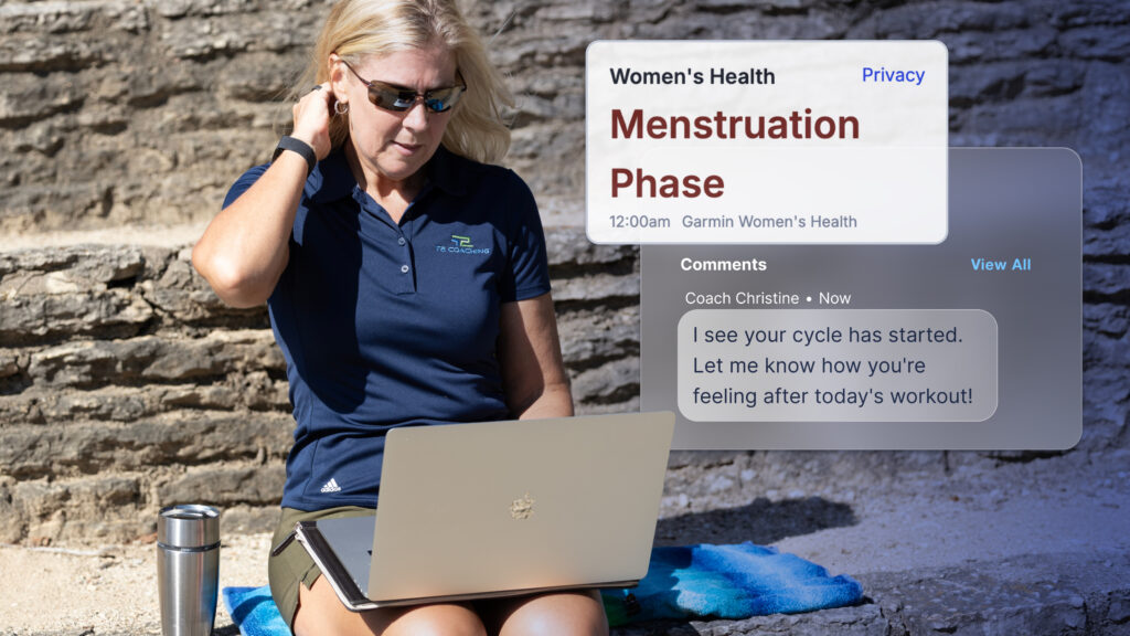 This image features a coach (blonde woman in sunglasses and a dark blue polo shirt) sitting outside, looking at her silver laptop. Overlaying the right side of the image is a screenshot of the Women’s Health Metric Card in TrainingPeaks, it’s titled: "Women's Health" with "Privacy" in the top right. Below this, it reads "Menstruation Phase" in large red font, with "12:00 am Garmin Women's Health" underneath. Further down, under "Comments," a message from "Coach Christine" (timestamped "Now") reads: "I see your cycle has started. Let me know how you're feeling after today's workout!" The overall scene suggests a coach monitoring an athlete's health data.