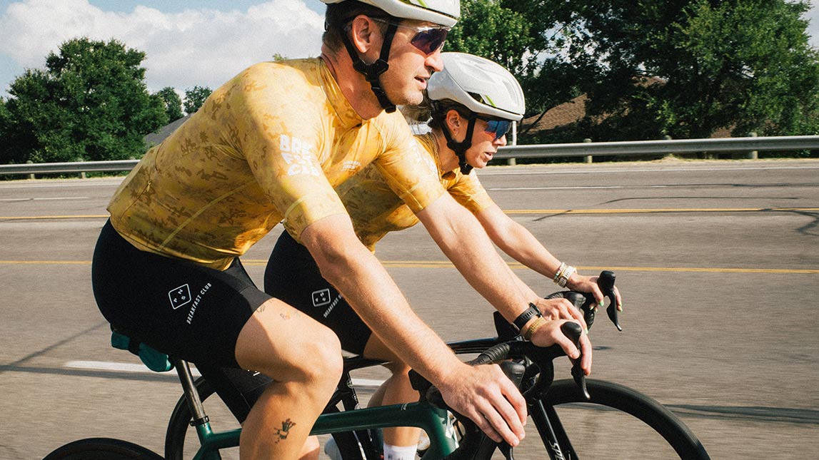 Pair Of Male Cyclists From Team Breakfast Club Riding Bikes On The Side Of The Road