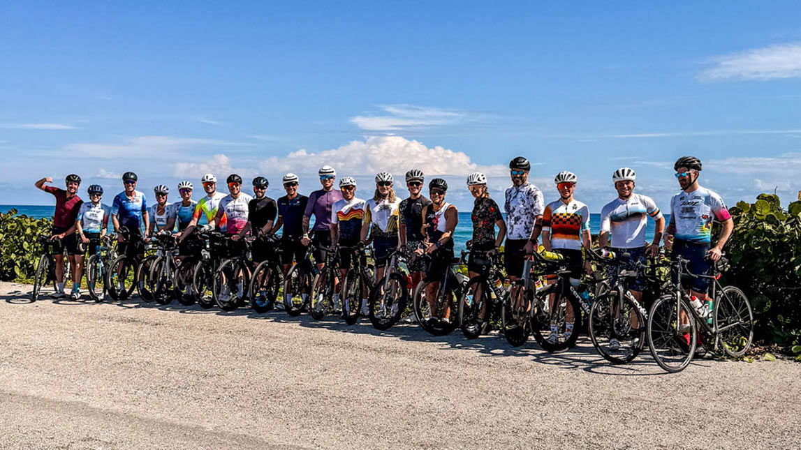 The Real Tri Squad Posing For Team Photo, Standing Next To Their Bikes On The Side Of The Road With The Ocean In The Background