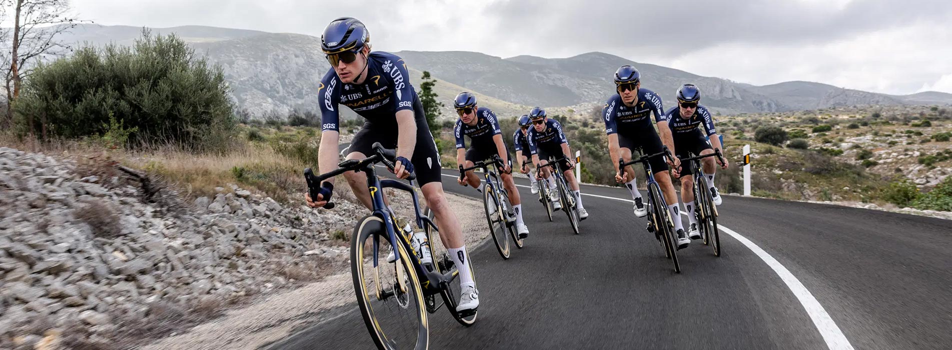 Male Cyclists From Team Pinarello Q36.5 Riding Down Winding Road With Mountains In The Background