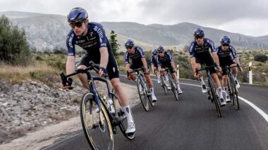 Male cyclists from team Pinarello-Q36.5 riding down winding road with mountains in the background