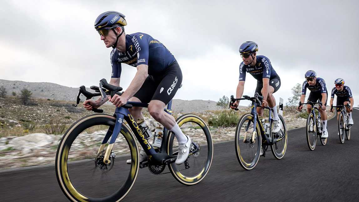 Male Cyclists From Team Pinarello Q36.5 Riding Down Smooth Paved Road With Mountains In The Background
